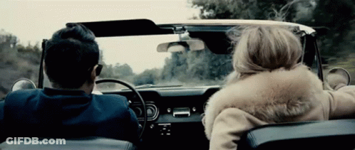 A Man And A Woman Are Driving In A Car With The Woman Wearing A Fur Coat GIF
