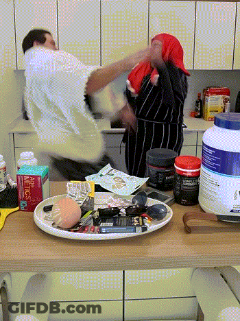 A Man And A Woman Are Fighting In A Kitchen With A Bottle Of Metcalf On The Counter Top GIF