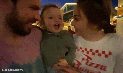 A Man And A Woman Are Holding A Baby . The Woman Is Wearing A Pizza Shirt . GIF