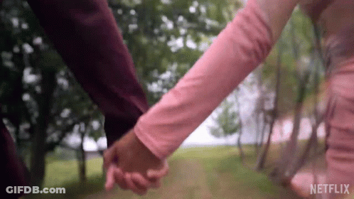 A Man And A Woman Are Holding Hands In A Park . The Woman Is Wearing A Pink Dress . GIF
