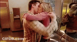 A Man And A Woman Are Hugging And Kissing In A Bathroom In Front Of A Mirror . GIF