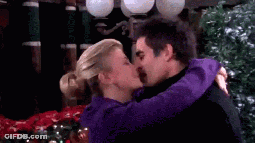 A Man And A Woman Are Kissing In Front Of A Christmas Tree . The Woman Is Wearing A Purple Jacket . GIF