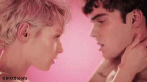 A Man And A Woman Are Kissing In Front Of A Pink Background . The Woman Is Touching The Man 'S Neck . GIF