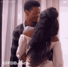 A Man And A Woman Are Kissing In Front Of A Window . The Woman Is Wearing A White Dress . GIF
