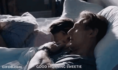 A Man And A Woman Are Laying In Bed And The Man Is Kissing The Woman On The Cheek . GIF
