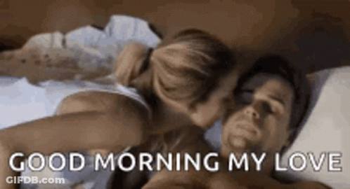 A Man And A Woman Are Laying In Bed And The Woman Is Kissing The Man On The Cheek . GIF