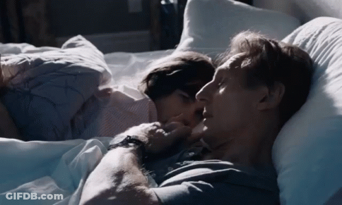 A Man And A Woman Are Laying In Bed And The Woman Is Kissing The Man 'S Neck GIF