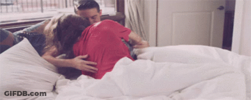 A Man And A Woman Are Laying On A Bed With White Sheets GIF