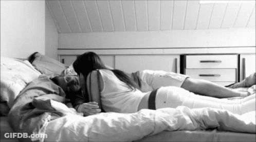 A Man And A Woman Are Laying On A Bed With White Sheets GIF