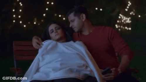 A Man And A Woman Are Laying On A Bench And The Man Is Holding A Phone GIF