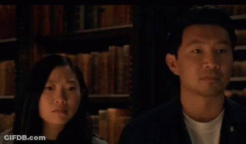 A Man And A Woman Are Looking At Each Other In Front Of A Bookshelf With The Words Or Written In Blue . GIF