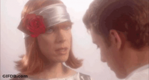 A Man And A Woman Are Looking At Each Other . The Woman Is Wearing A Headband With A Rose In Her Hair . GIF