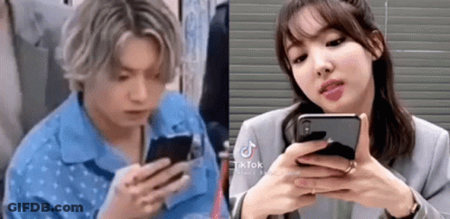A Man And A Woman Are Looking At Their Phones . The Man Is Wearing A Blue Shirt . GIF