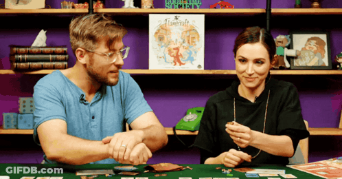 A Man And A Woman Are Playing A Board Game With A Sign In The Background That Says ' Roguelike Society ' GIF