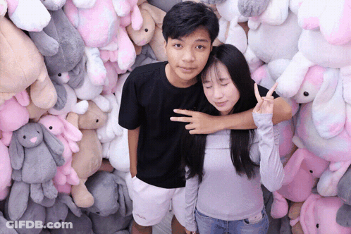 A Man And A Woman Are Posing For A Picture In Front Of A Wall Of Stuffed Animals GIF