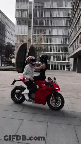 A Man And A Woman Are Riding A Red Motorcycle In Front Of A Building GIF