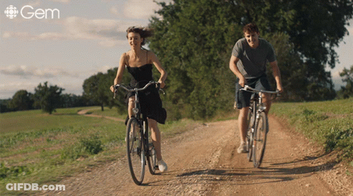 A Man And A Woman Are Riding Bicycles Down A Dirt Road And The Word Gem Is On The Bottom Right GIF
