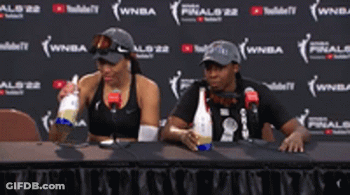 A Man And A Woman Are Sitting At A Table Drinking From Bottles GIF