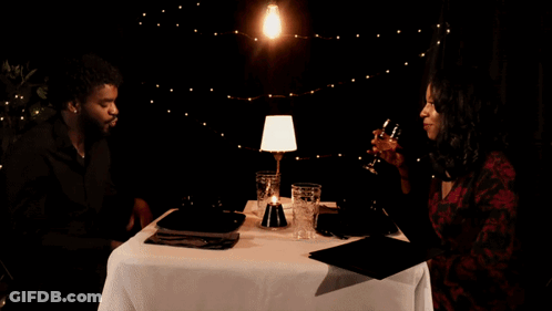 A Man And A Woman Are Sitting At A Table Drinking Wine And Talking GIF