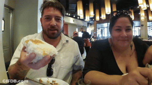 A Man And A Woman Are Sitting At A Table Eating Food GIF