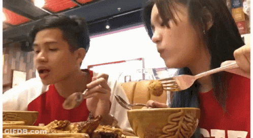 A Man And A Woman Are Sitting At A Table Eating Food With Spoons And Forks . GIF
