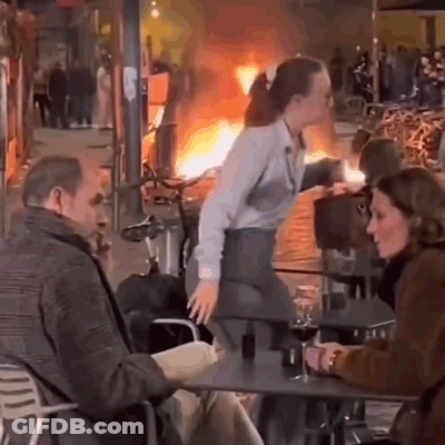 A Man And A Woman Are Sitting At A Table In Front Of A Burning Building . GIF