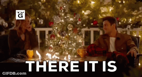 A Man And A Woman Are Sitting At A Table In Front Of A Christmas Tree With The Words There It Is Above Them . GIF
