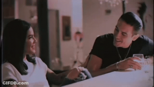 A Man And A Woman Are Sitting At A Table Talking To Each Other And Smiling . GIF