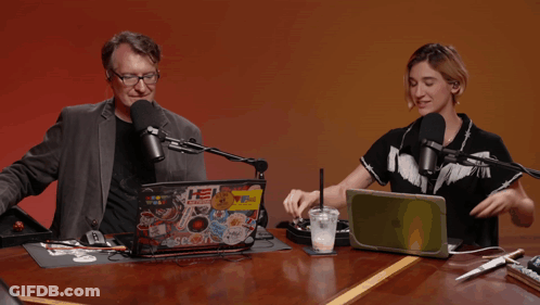 A Man And A Woman Are Sitting At A Table With A Laptop With Stickers On It That Says He Radio GIF