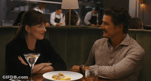 A Man And A Woman Are Sitting At A Table With A Martini GIF