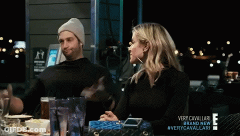 A Man And A Woman Are Sitting At A Table With A Sign That Says Very Cavallari On It GIF