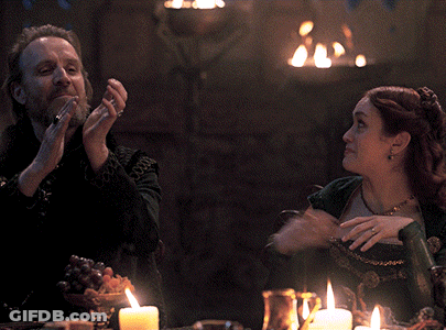 A Man And A Woman Are Sitting At A Table With Candles And A Fire In The Background GIF