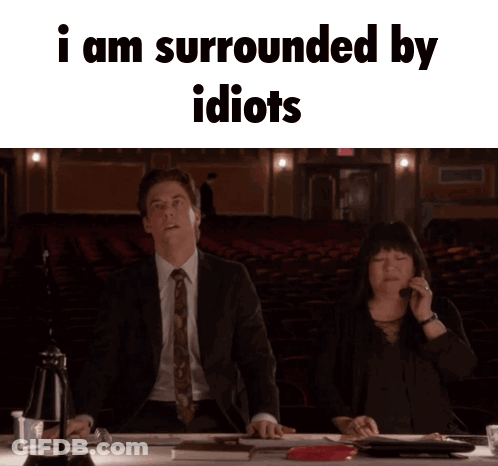 A Man And A Woman Are Sitting At A Table With The Words I Am Surrounded By Idiots GIF
