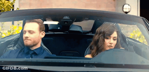 A Man And A Woman Are Sitting In A Car Looking Out The Window GIF