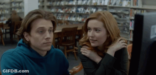 A Man And A Woman Are Sitting In A Library Looking At A Computer Screen GIF