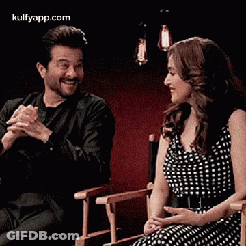 A Man And A Woman Are Sitting In Chairs With Their Hands Folded . GIF