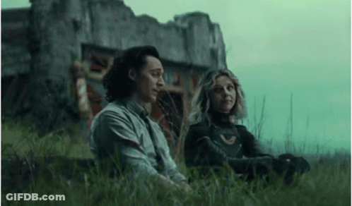 A Man And A Woman Are Sitting In The Grass Next To A Building . GIF