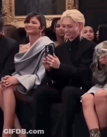 A Man And A Woman Are Sitting Next To Each Other At A Fashion Show . The Woman Is Holding A Cell Phone . GIF
