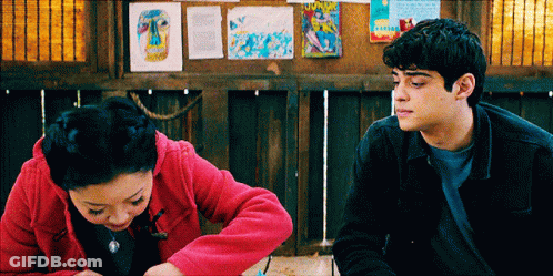 A Man And A Woman Are Sitting Next To Each Other In Front Of A Bulletin Board GIF