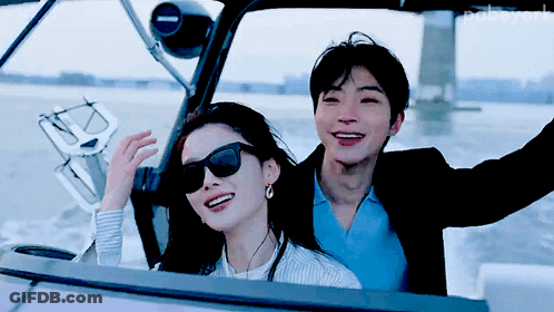 A Man And A Woman Are Sitting On A Boat And The Man Is Wearing Sunglasses And The Woman Is Wearing Earrings GIF