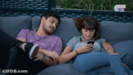 A Man And A Woman Are Sitting On A Couch And Looking At Their Phones . GIF