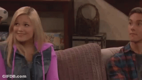 A Man And A Woman Are Sitting On A Couch And Smiling . The Woman Is Wearing A Pink Hoodie . GIF