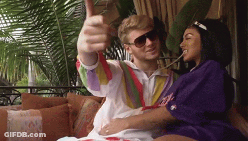 A Man And A Woman Are Sitting On A Couch And The Man Is Wearing Sunglasses And Giving A Thumbs Up GIF
