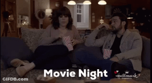 A Man And A Woman Are Sitting On A Couch Eating Popcorn And Watching A Hallmark Movie GIF