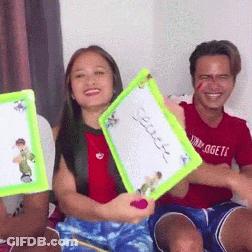 A Man And A Woman Are Sitting On A Couch Holding A White Board With A Picture On It . GIF