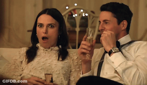 A Man And A Woman Are Sitting On A Couch Holding Glasses Of Champagne GIF
