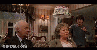 A Man And A Woman Are Sitting On A Couch In A Living Room With A Cake On Their Head . GIF