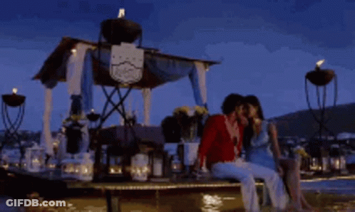 A Man And A Woman Are Sitting On A Dock Kissing Each Other GIF