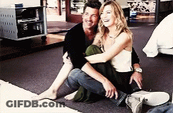 A Man And A Woman Are Sitting On The Floor Hugging Each Other And Smiling . GIF