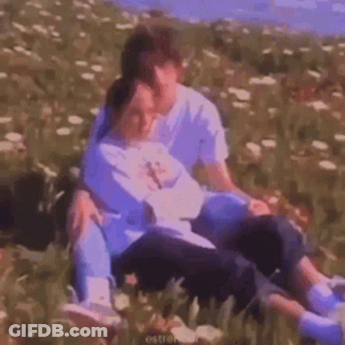 A Man And A Woman Are Sitting On Top Of Each Other In A Field Of Flowers . GIF